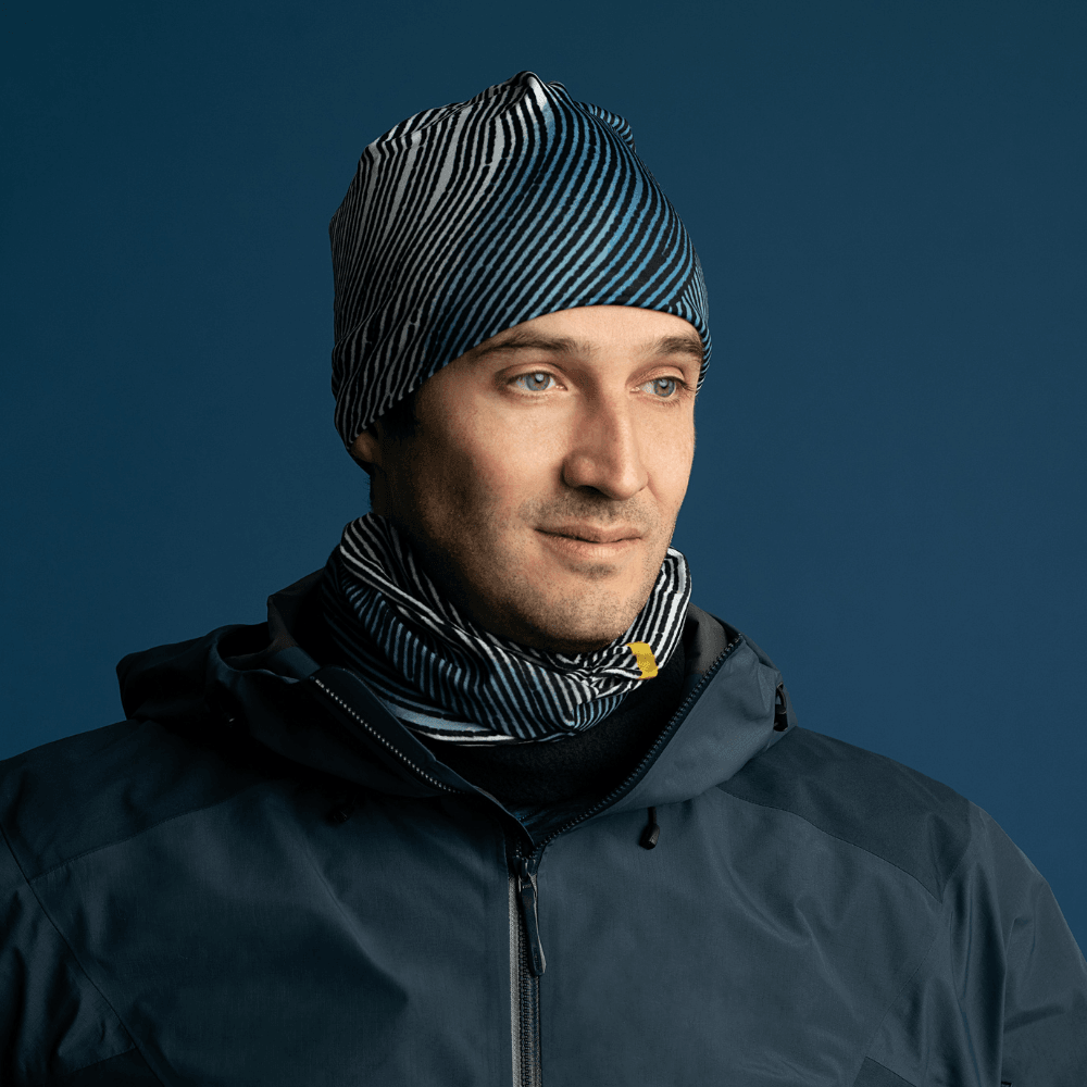 A man wearing a dark winter jacket and a matching beanie, with the Abstract Lines multifunctional tube worn as a neck gaiter, looking to the side.