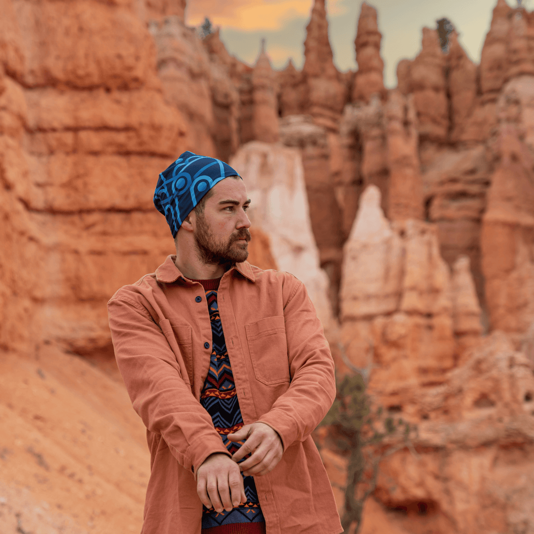 Homme barbu portant une chemise saumon et une tuque à motif, debout dans un paysage désertique.