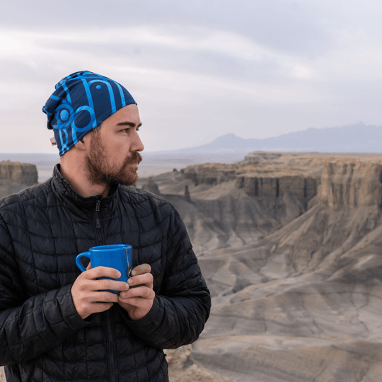 Homme portant une veste noire matelassée et une tuque à motif bleu, tenant une tasse bleue dans un paysage rocheux.