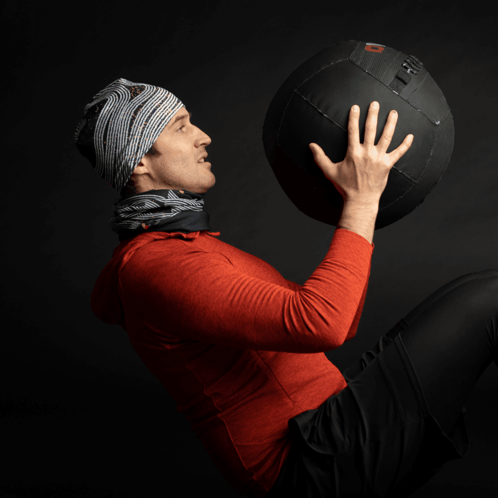 Man in red hoodie and black shorts wearing the Dark Steel Multifunctional Tube as a neck warmer while holding a medicine ball