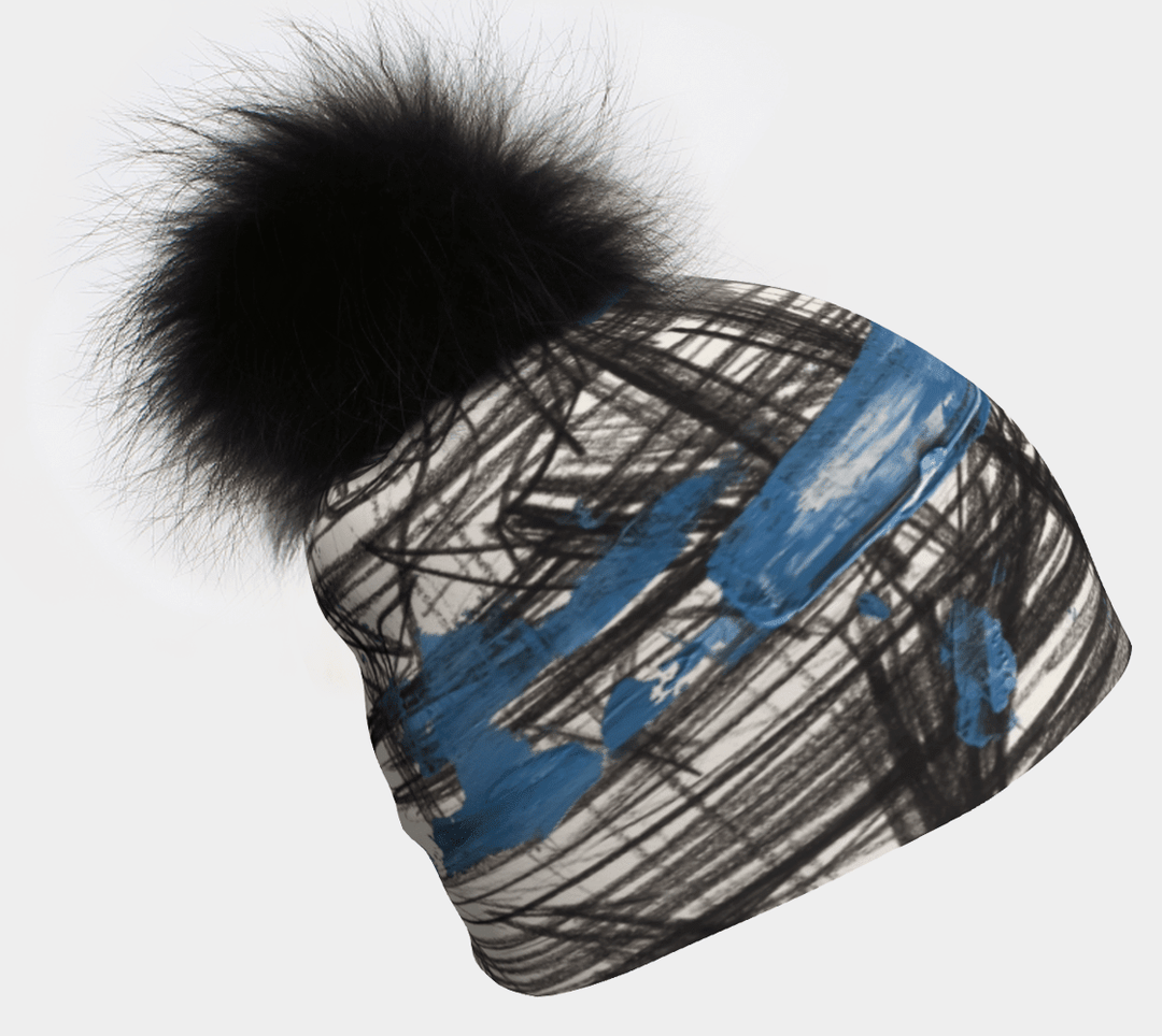 A close-up of a white beanie with an abstract black sketch pattern and bold blue strokes, topped with a fluffy black pompom.