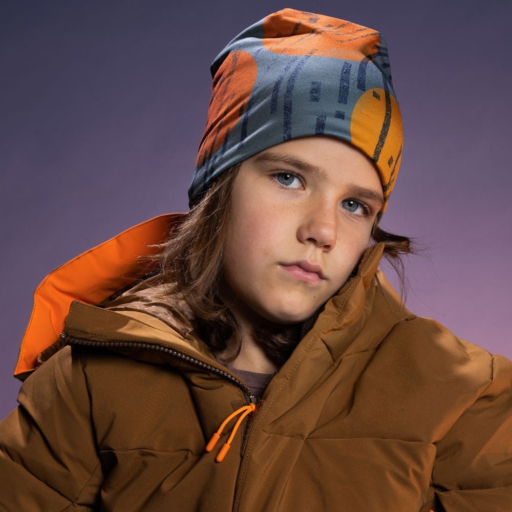 Enfant portant la tuque artistique orange et grise avec un manteau matelassé brun, fond violet en studio