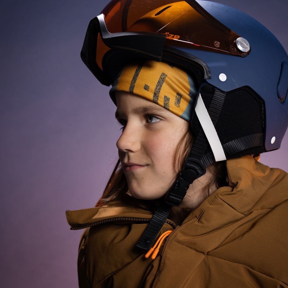 Jeune skieur portant la tuque Orange Movement colorée sous un casque de ski marine avec lunettes orange, lumière violet studio