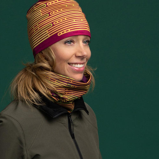 Femme souriant portant une tuque et un cache-cou magenta assortis, avec une veste verte.