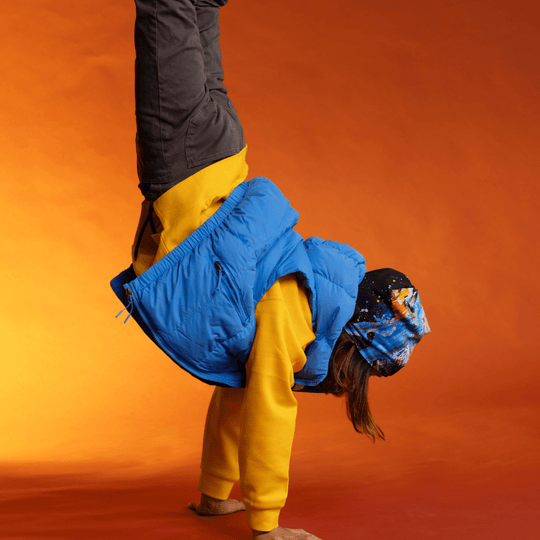 Une personne en gilet bleu et sweat à capuche jaune effectuant un poirier, portant la tuque MatriX.