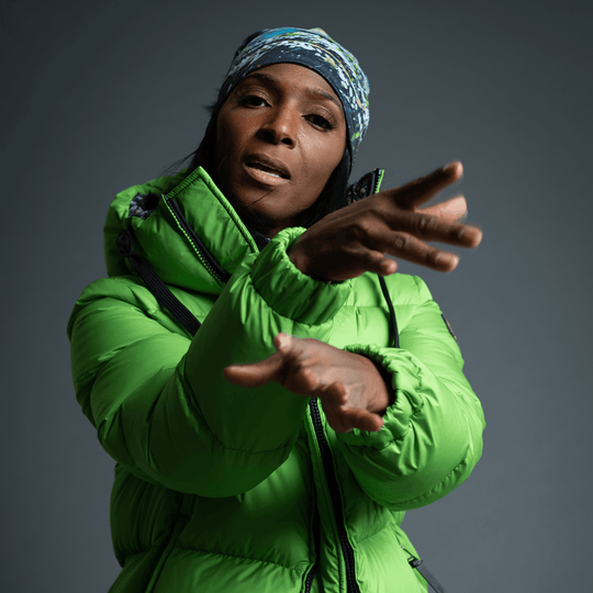 Femme en veste matelassée verte, faisant un geste, portant la tuque Méduse en bandeau.