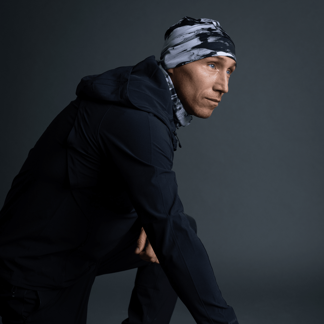 Homme en veste noire, tuque et cache-cou au motif abstrait noir et blanc, en pose accroupie.