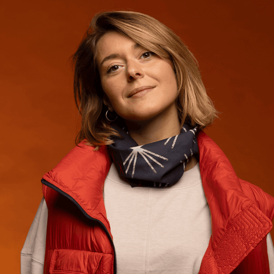 Woman wearing a red puffer vest and white sweater, with the MotifX tube styled as a neck gaiter displaying a dark blue background with a white abstract starburst pattern.
