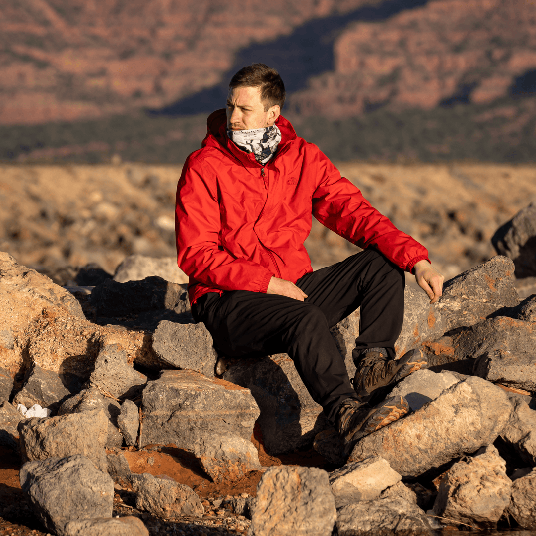 Man in a red jacket sitting on rocks outdoors, wearing the black and white abstract pattern neck gaiter.