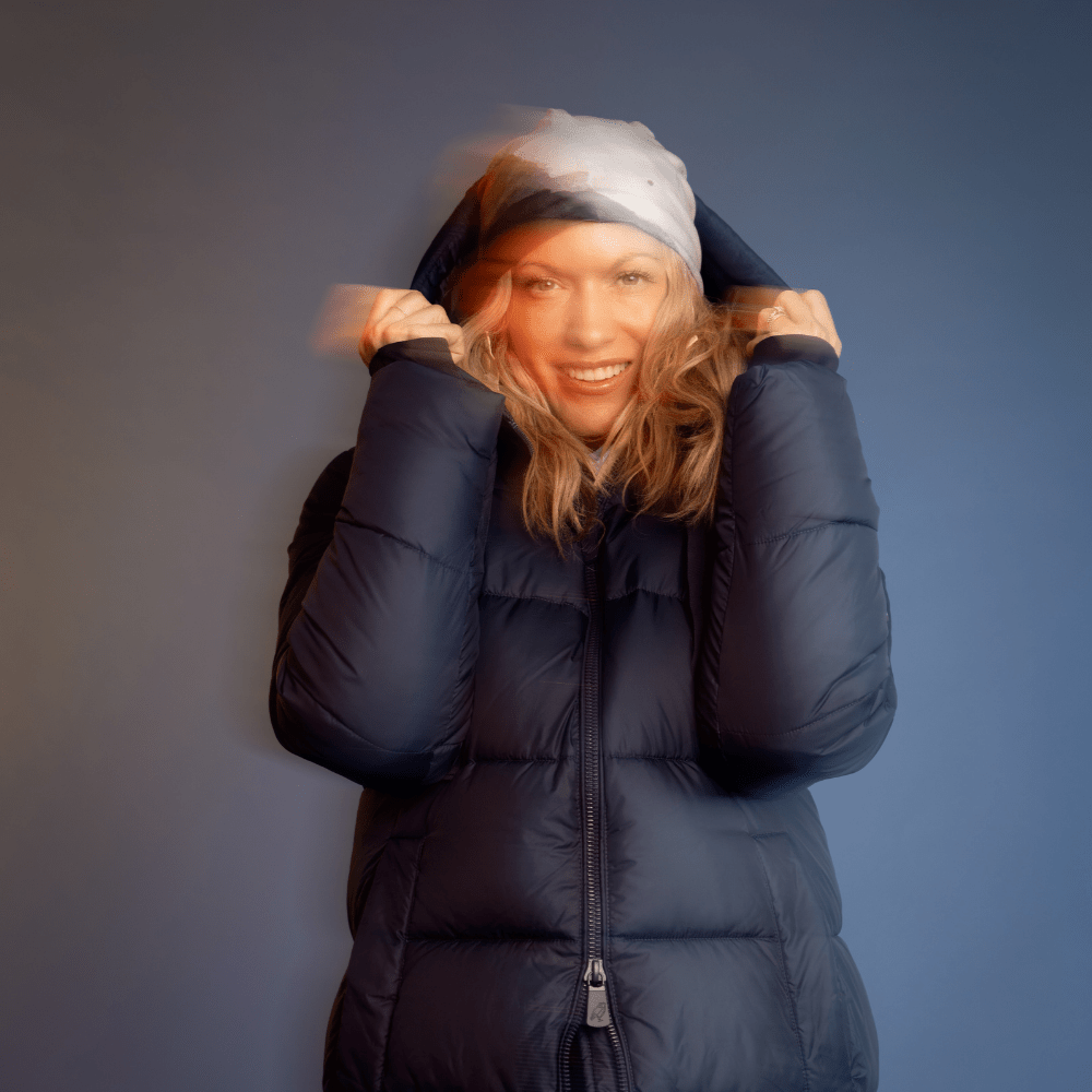Woman in a puffer jacket pulling up the hood of her beanie, smiling.