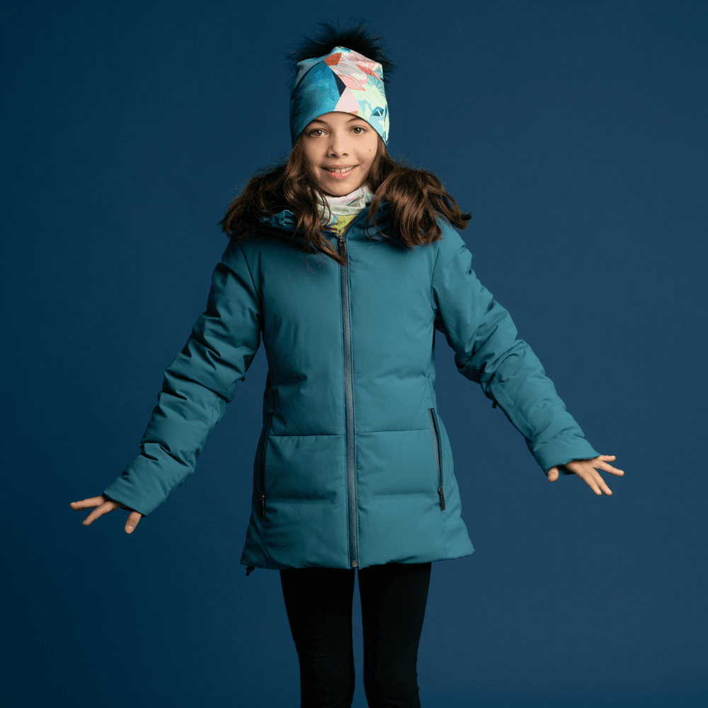 Une jeune fille portant une veste d'hiver turquoise, une tuque à motif assorti et un cache-cou, souriant.