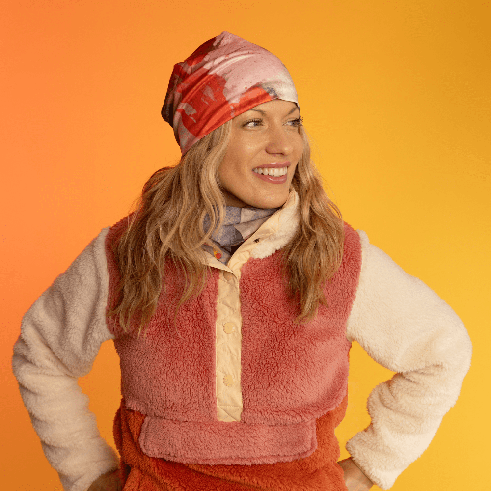 Woman wearing a beanie with pink and red abstract art, smiling against an orange background.