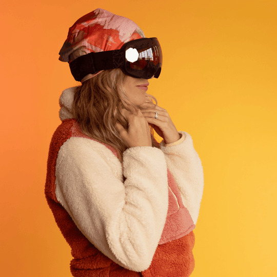 Woman wearing a beanie with abstract art and ski goggles, against an orange background.