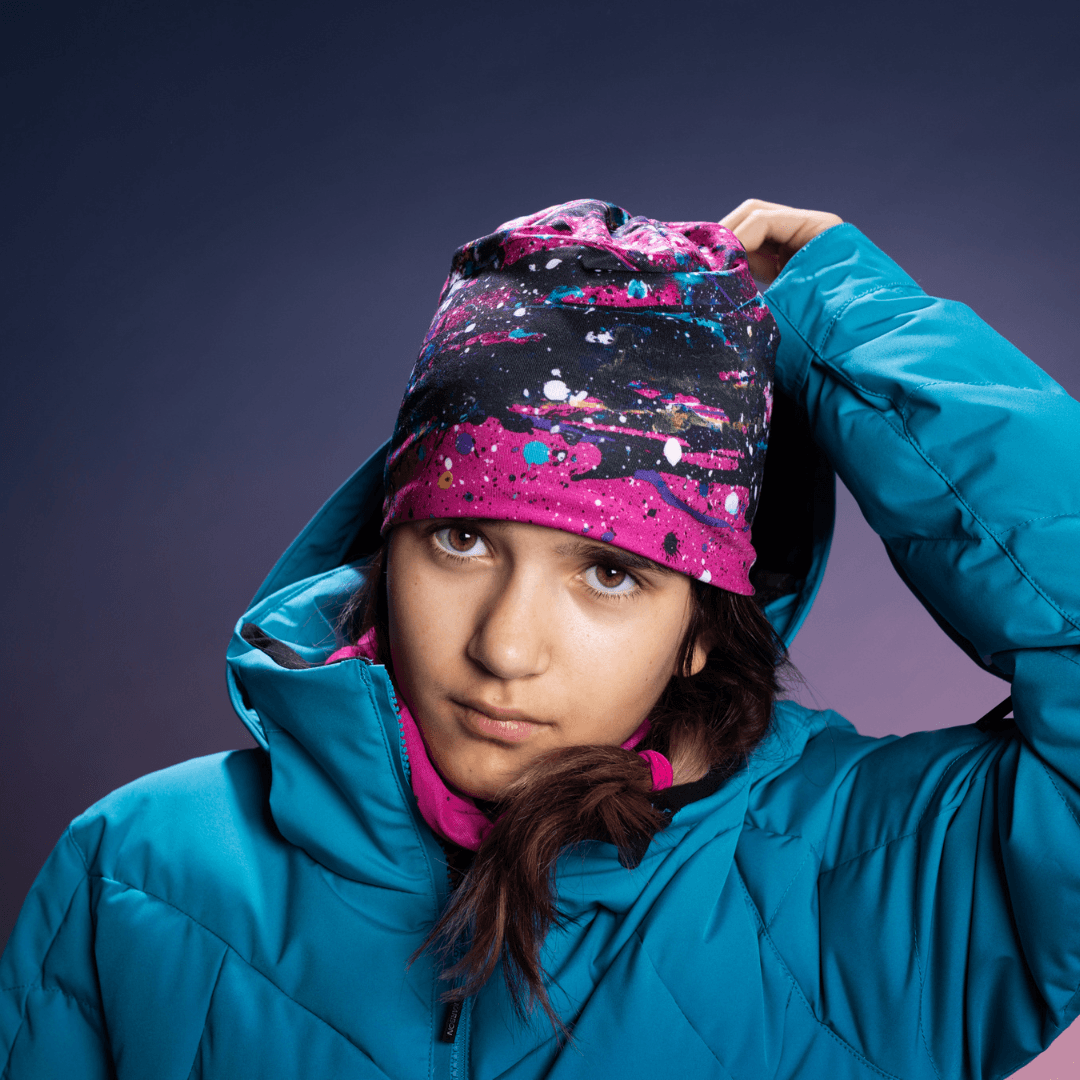 Jeune fille en veste de ski bleue portant une tuque rose à imprimé abstrait et un cache-cou rose, regardant la caméra.