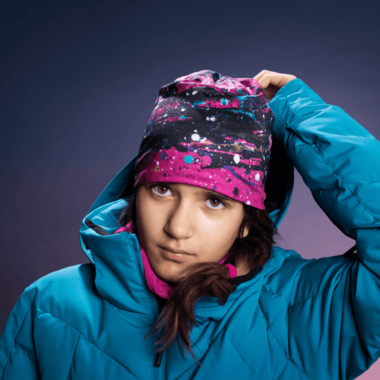 Jeune fille en veste de ski bleue portant une tuque rose à imprimé abstrait et un cache-cou rose, regardant la caméra.