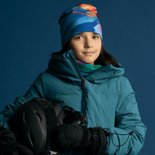 A young girl in a teal winter jacket holding a ski helmet, wearing a colorful neck gaiter and matching beanie.