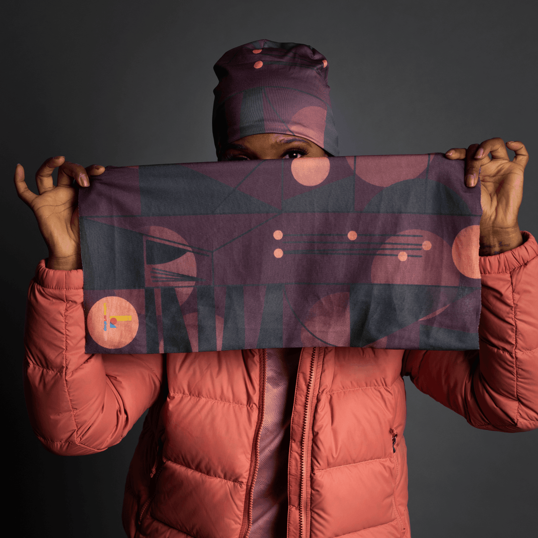 A person wearing a coral puffer jacket holds up a rectangular fabric tube with a geometric purple and dark grey pattern, their face partially obscured.