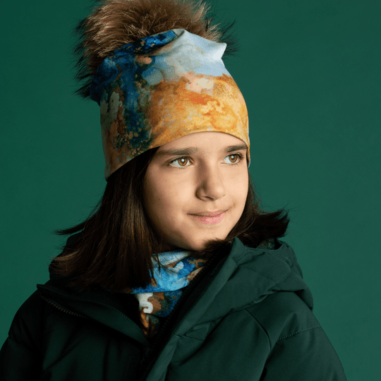 A young girl wearing a green winter jacket and a POLAR Ocean Marble beanie with a pompom, with the matching neck warmer visible.