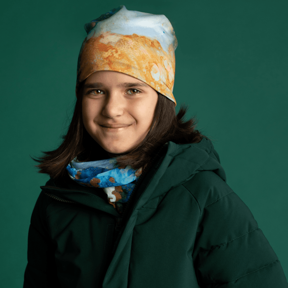 A young girl wearing a green winter jacket, a POLAR Ocean Marble beanie, and the matching neck warmer.