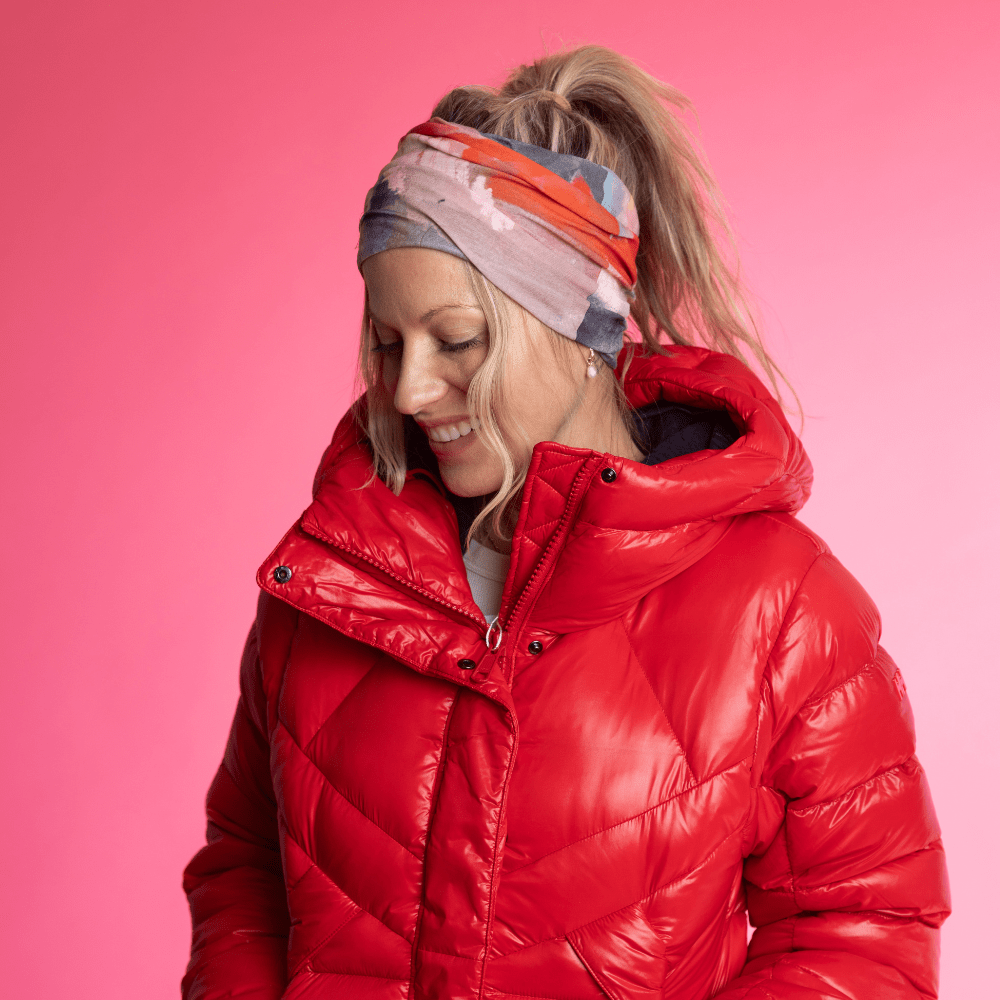 Woman wearing the POLAR Pink &amp; Flames Multifunctional Tube as a headband, looking down with a smile, styled with a red puffer jacket against a gradient pink background.