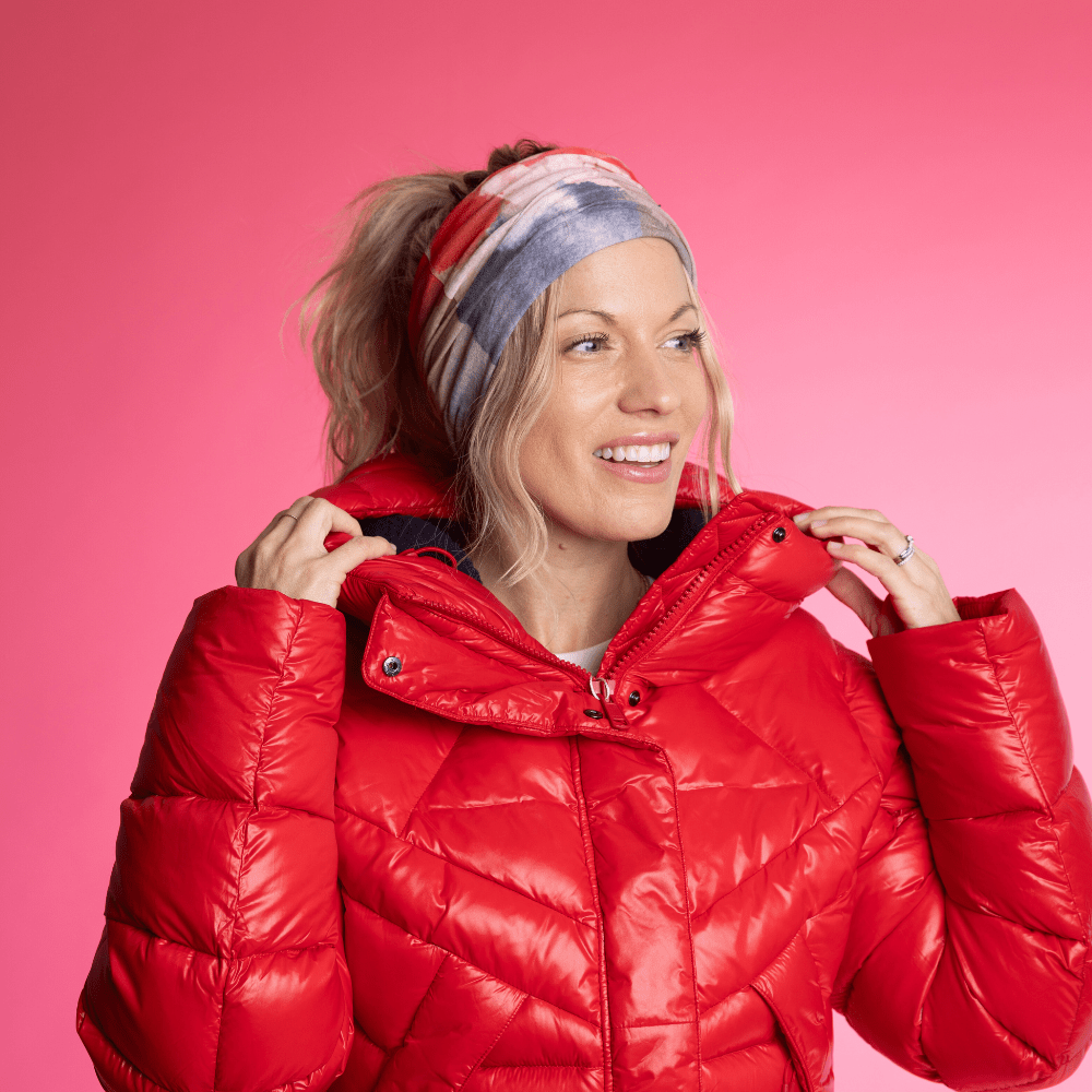 Woman wearing the POLAR Pink &amp; Flames Multifunctional Tube as a headband, styled with a red puffer jacket against a bright pink background.