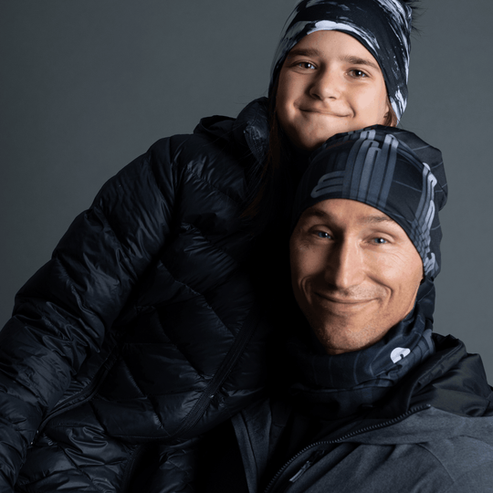 A man and a child wearing matching black beanies and neck tubes with a grey graphic design, bundled in winter jackets.
