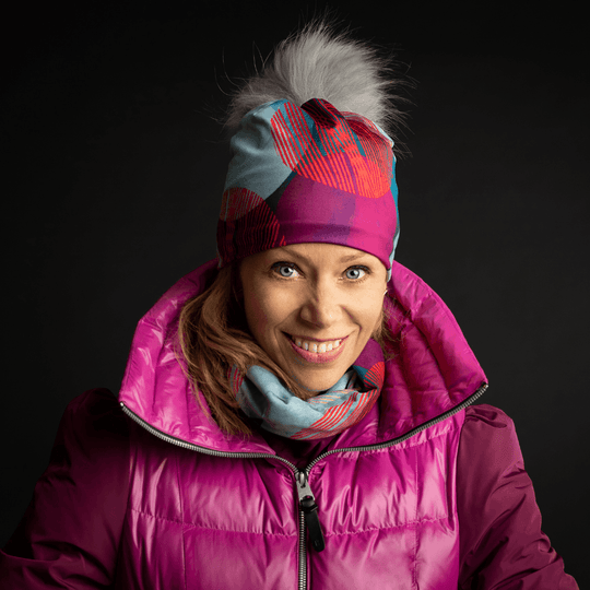 A smiling woman wearing a pink puffer vest and a matching Teal Dots beanie and neck gaiter.