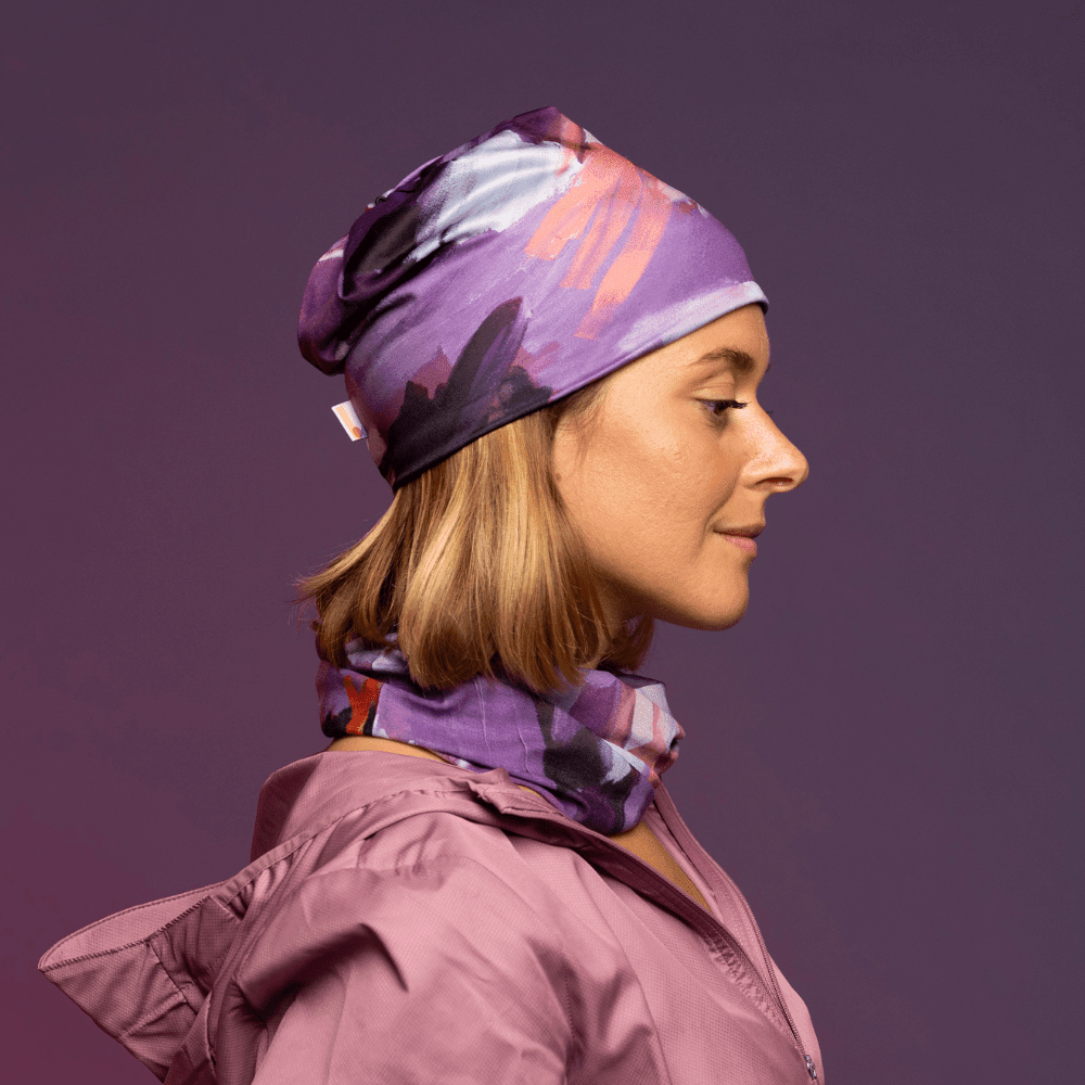 A woman wearing a purple, pink, and white abstract print beanie and neck tube over a pink jacket.