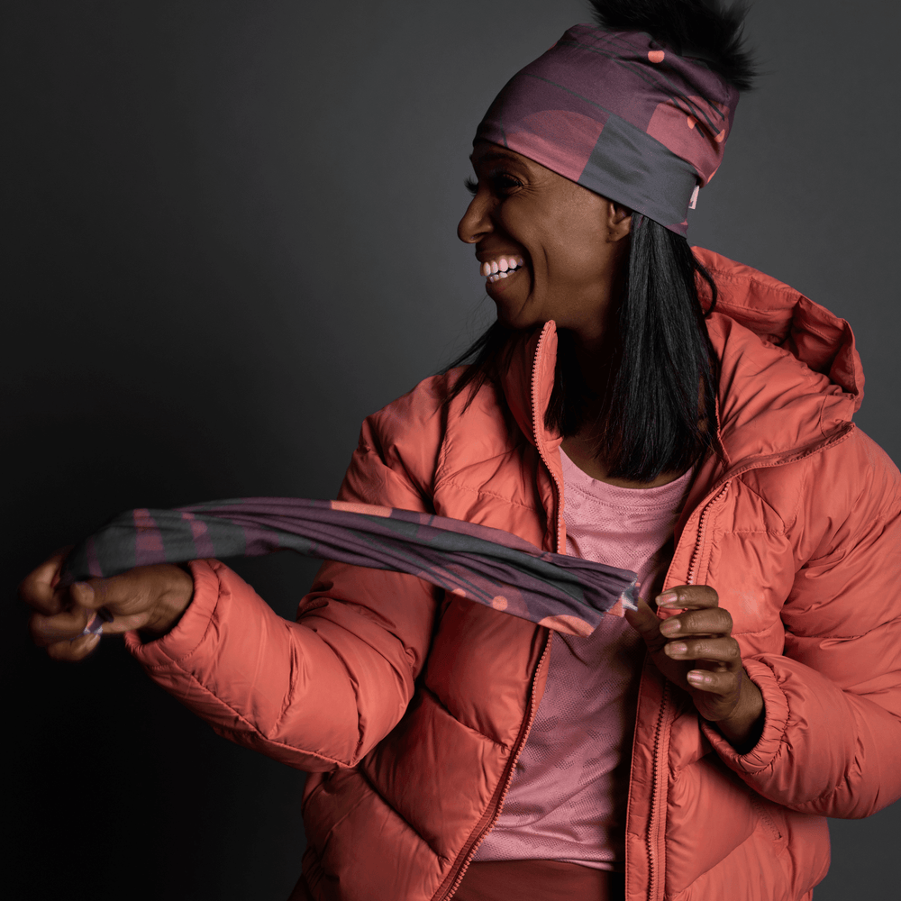 Une femme portant une veste d'hiver corail tient un cache-cou assorti à motifs géométriques violets, souriant.