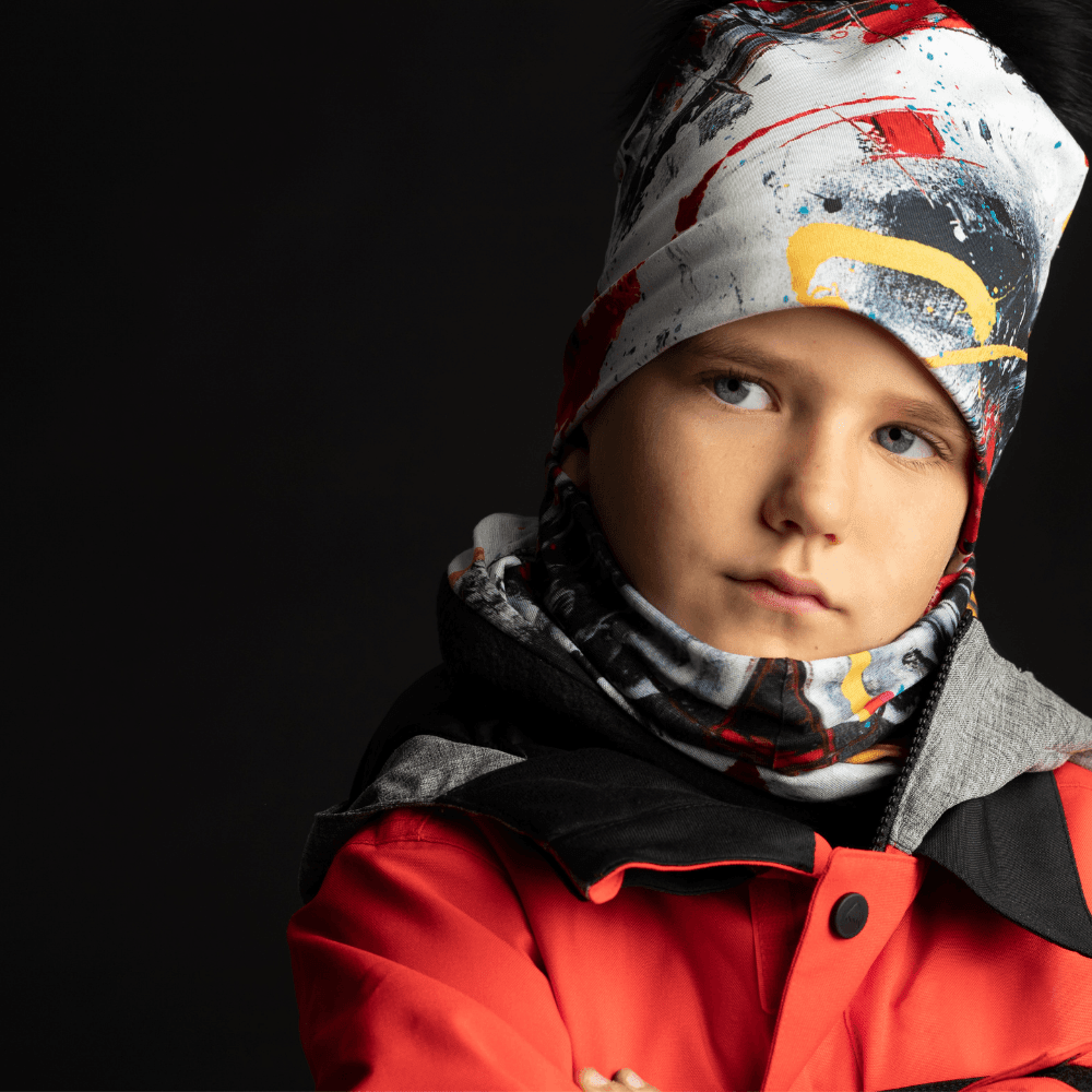 Close-up of a young boy wearing a ski jacket, with a matching beanie and neck tube featuring abstract red, black, and yellow paint splatters.