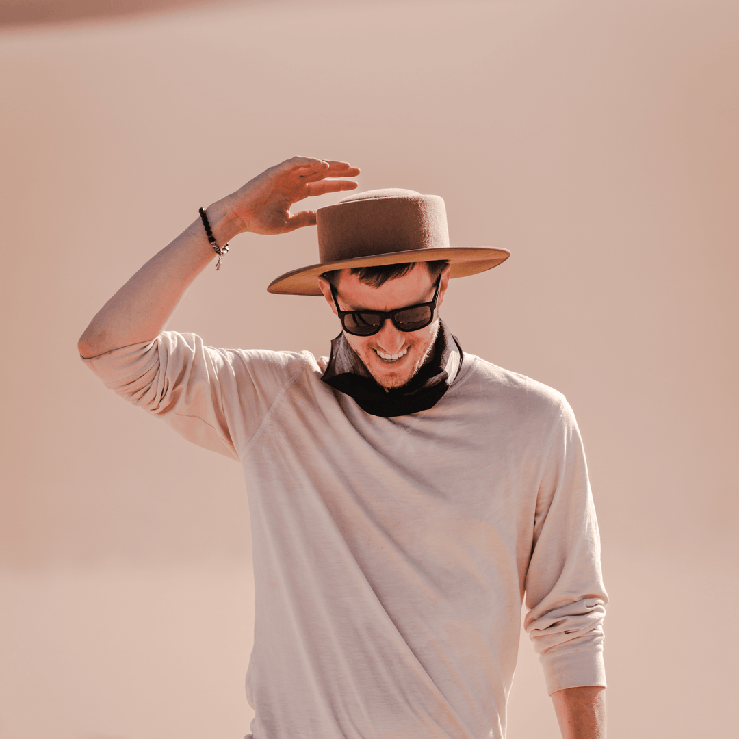 Man wearing a tan fedora and sunglasses, tipping his hat with one hand, with a black multifunctional neck tube casually worn around his neck.