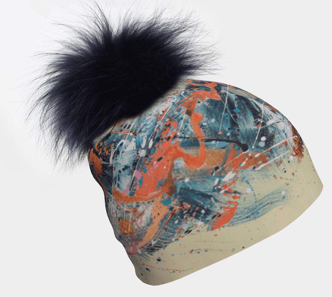 A close-up view of a Sphénédon Beanie with a navy pompom, showcasing abstract colourful paint splatter details on a cream background.