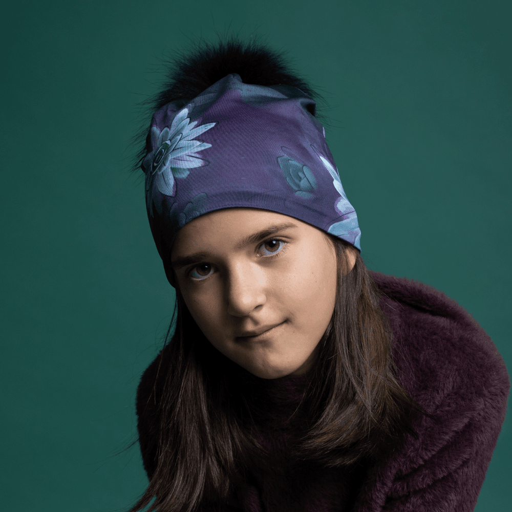 Jeune femme portant une tuque à imprimé succulent violet avec pompon en fourrure noire, stylisée avec un manteau prune sur fond sarcelle