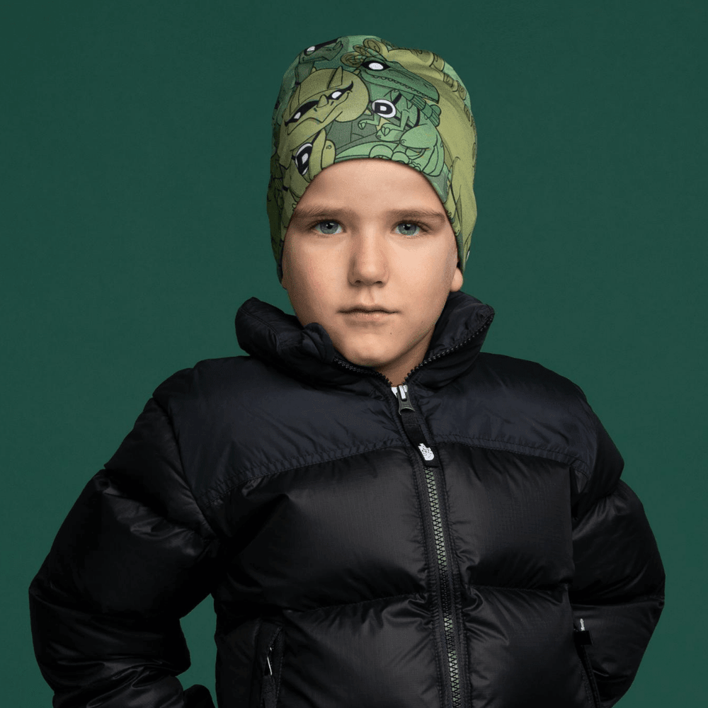 Un jeune garçon portant une tuque verte avec un imprimé de dinosaure, vêtu d'une veste noire par-dessus un fond vert foncé.