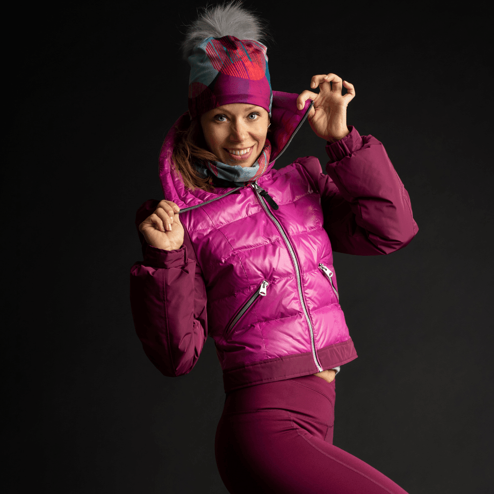 Une femme en tenue de sport rose sourit, ajustant sa tuque Pois-Bleu avec pompon gris détachable et sa veste rose.