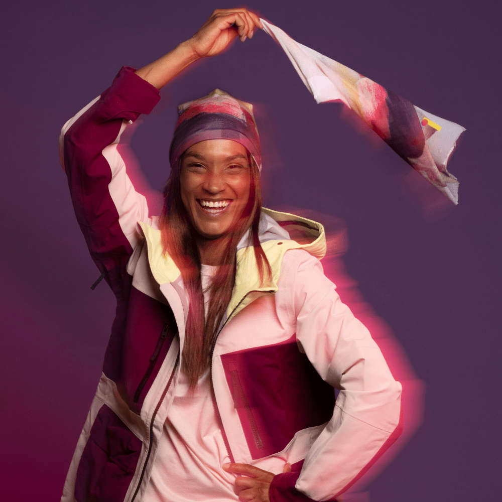 Une femme souriante portant la tuque "Le Nomade Artistique" et une veste de ski colorée, tenant une écharpe assortie.