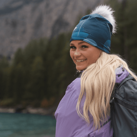 Femme souriante aux cheveux blonds portant la tuque bleu-vert avec un pompon gris et une veste violette, regardant par-dessus son épaule.
