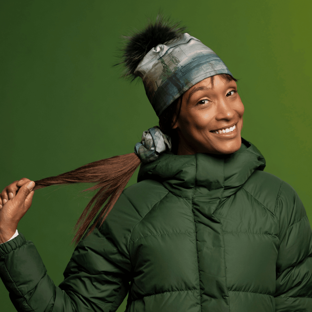 Une femme souriante portant un manteau d'hiver vert et la Tuque Murale à Pompon tire sur sa queue de cheval.