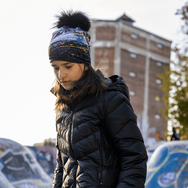 Young girl in profile, looking down and looking at the Abracadabra hat with detachable pompom illustrated by Mégane Fortin. This hat with abstract patterns is made of bamboo to wick away moisture. Perfect hat for hiking, walking, and outdoor activities. 