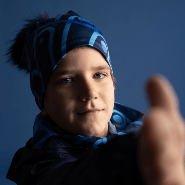 Boy reaching out, wearing the mechanic beanie hat designed by Zaire. The beanie is in blue tones with geometric shapes and a black pom-pom.