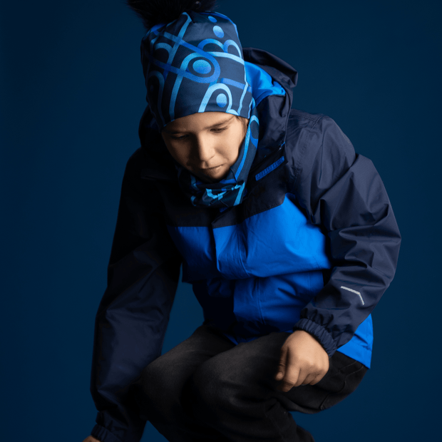 Boy jumping, wearing the mechanic beanie hat designed by Zaire. The bamboo beanie with removable pom is in blue tones with geometric shapes and a black pom-pom. Perfect gift for men and boys